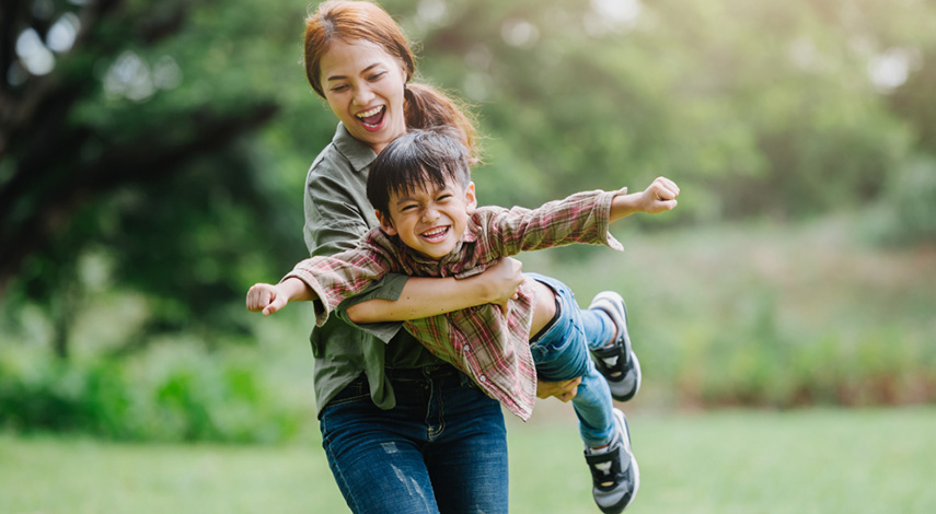 A woman holds a small child in her arms as he pretends to be a flying super hero.