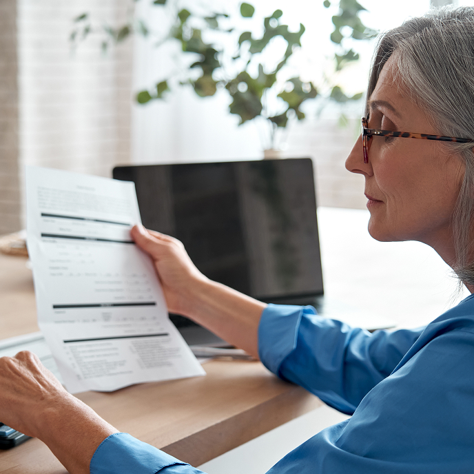 Older woman looking at a financial document at a desk. There’s a laptop on the desk next to her.
