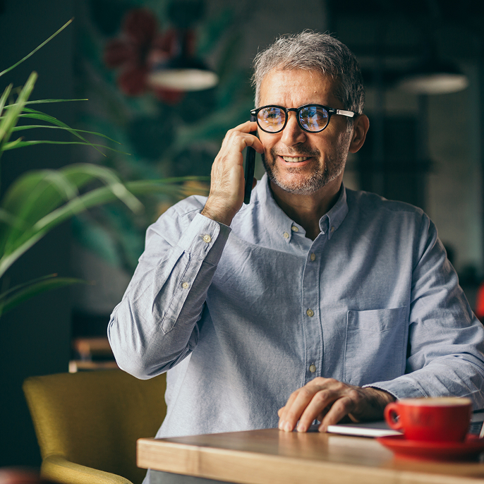 Man is sitting at a table and talking on a cell phone. There’s a laptop and a red cup and saucer in front of him on the table.