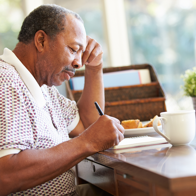 A retired man is sitting at a table writing in a notebook. He has a coffee mug and a muffin on a plate in front of him.