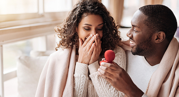 A young couple is shown as the man proposes to the woman. Grinning, he holds open a small red box with a ring inside. She clasps her hands over her mouth in delight.