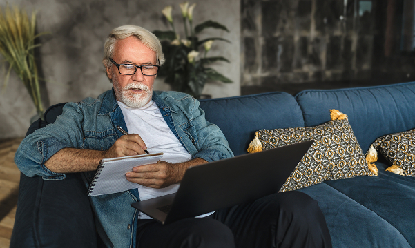 A man in his mid-50s sits on a couch while taking notes about preparing for retirement in a notebook with a laptop on his lap.